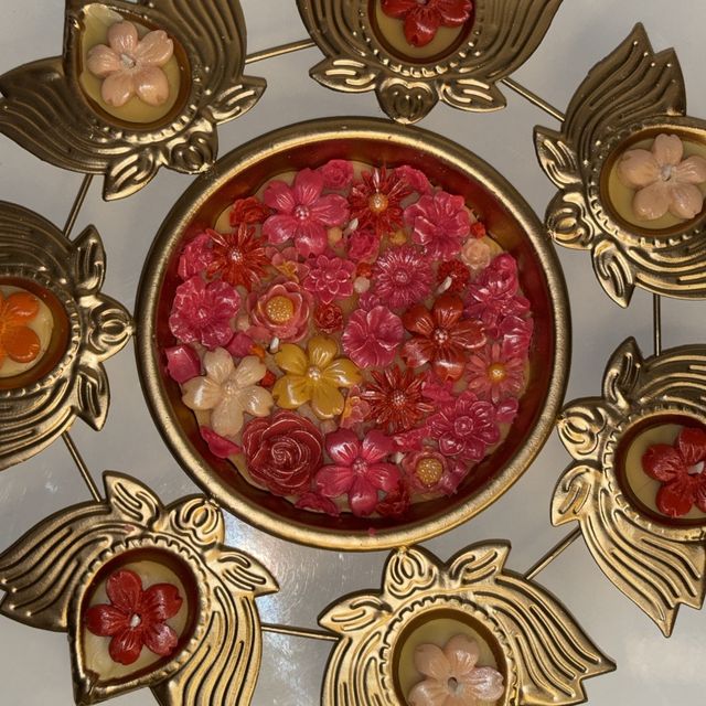 DH-01: Seven lotus diyas on a gold metal stand, around an urli of pink and red blooms, close-up of the flower bowl.