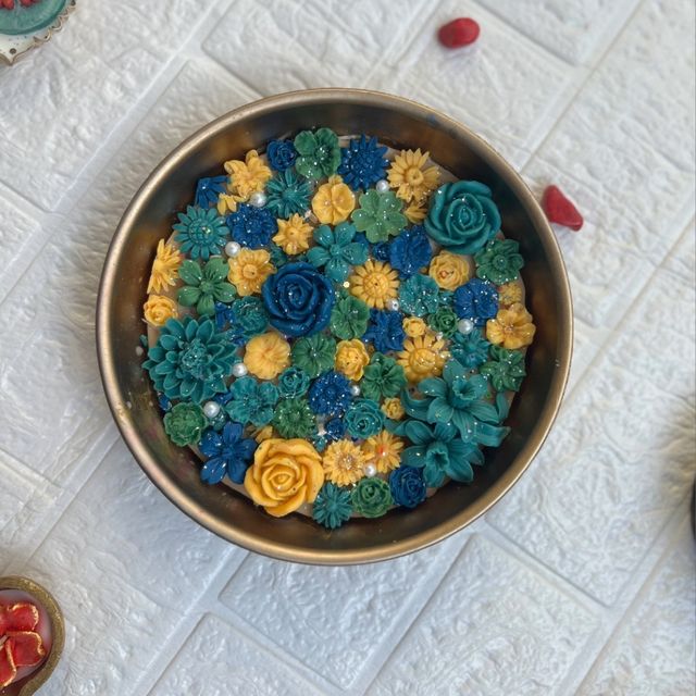 DH-05: An antique-gold metal urli full of blue, teal and yellow flowers, on a white table.